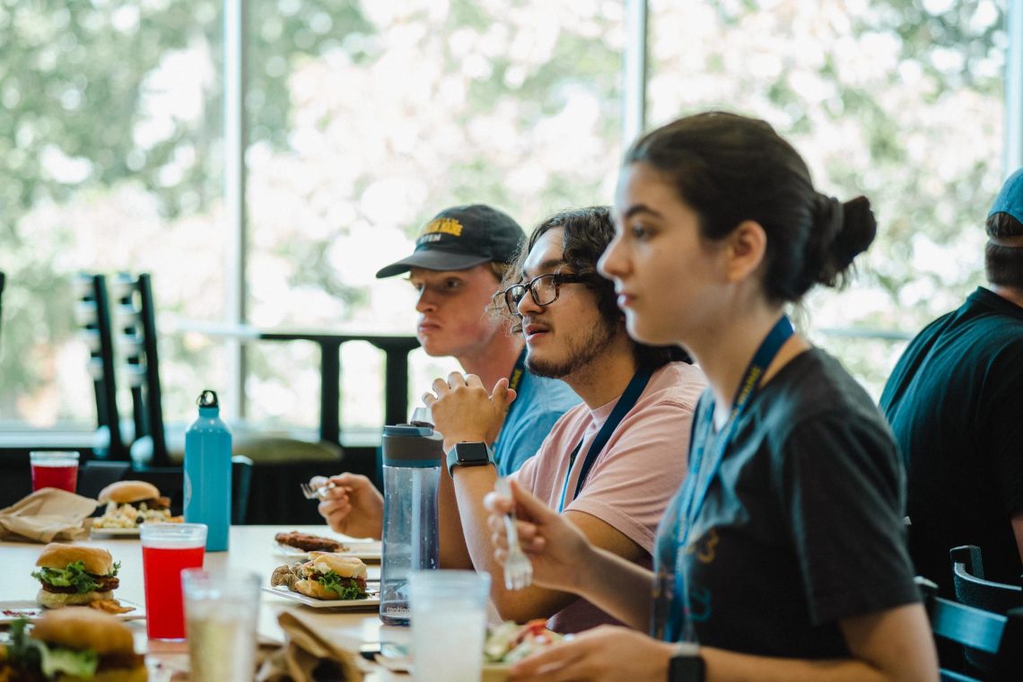 Students eat lunch together at a table while listening attentively to something off-camera during a campus event.