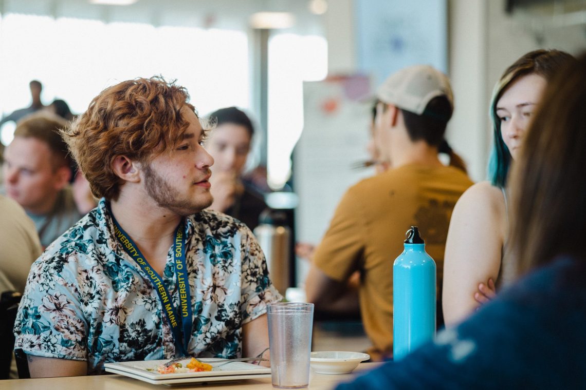 A student wearing a University of Southern Maine lanyard talks with others at a table in a busy campus dining hall.
