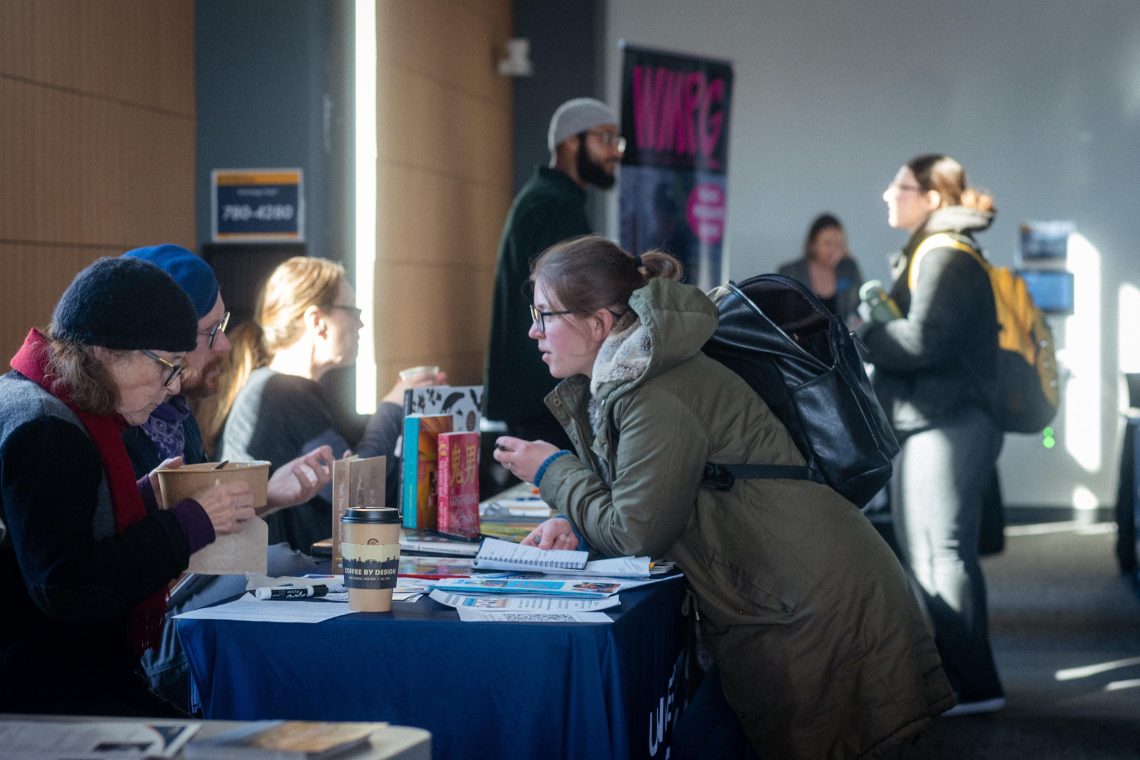 People chat and browse books and flyers at a table event indoors, with a "Coffee by Design" cup and a WMPG banner visible nearby.