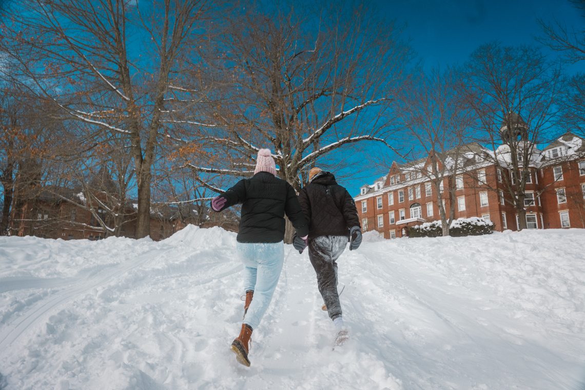Two students in winter coats and boots run hand-in-hand across a snow-covered path toward a brick dormitory on campus