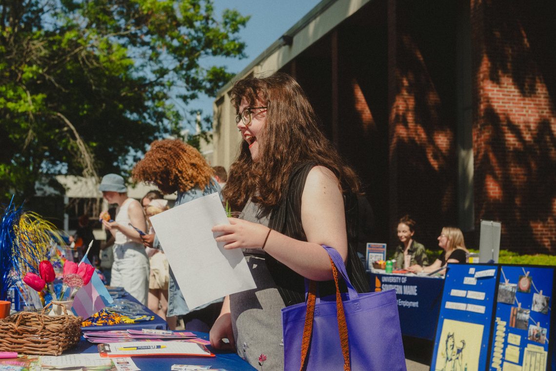 A student laughs while looking at a flyer at an outdoor campus event, with tables of activities and a University of Southern Maine banner in the background.