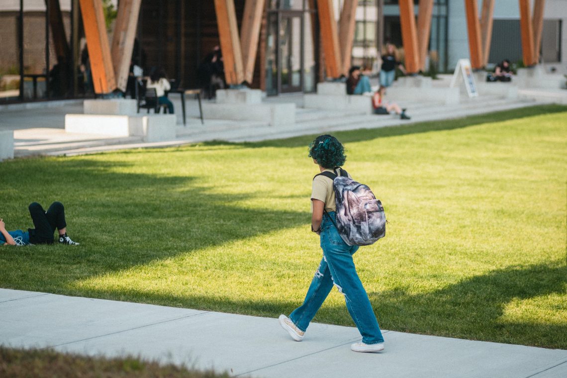 A student with teal hair and a backpack walks along a sunny sidewalk at USM's Gorham campus, with other students lounging on the grass and the Wishcamper Center's distinctive wooden columns visible in the background.