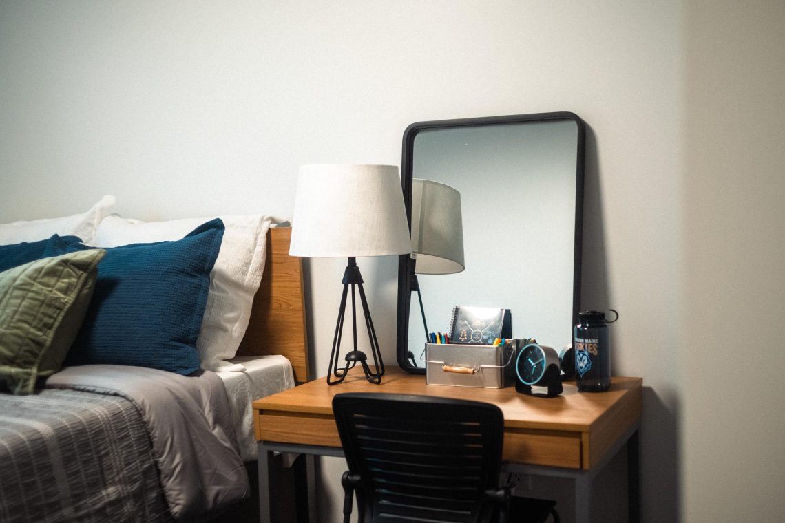 Desk lamp, mirror and clock arranged on a wooden desk beside a bed with blue and green pillows in a dorm room