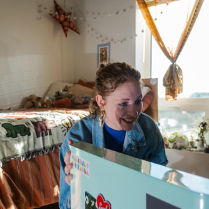 Student smiles while opening a mini fridge covered in stickers in a decorated dorm room with string lights and a patterned blanket