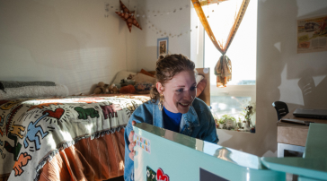 Student smiles while opening a mini fridge covered in stickers in a decorated dorm room with string lights and a patterned blanket