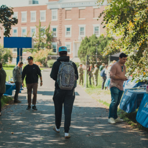 Students walk along a tree-lined pathway between rows of blue-draped resource and club tables during an outdoor fair on USM's Portland campus, with a brick academic building visible in the background.