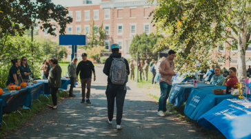 Students walk along a tree-lined pathway between rows of blue-draped resource and club tables during an outdoor fair on USM's Portland campus, with a brick academic building visible in the background.