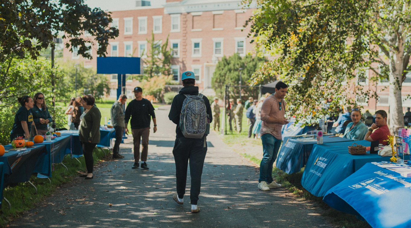 Students walk along a tree-lined pathway between rows of blue-draped resource and club tables during an outdoor fair on USM's Portland campus, with a brick academic building visible in the background.