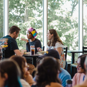 Students chat over lunch at a table by large windows in a busy campus dining hall.