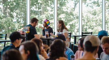 Students chat over lunch at a table by large windows in a busy campus dining hall.