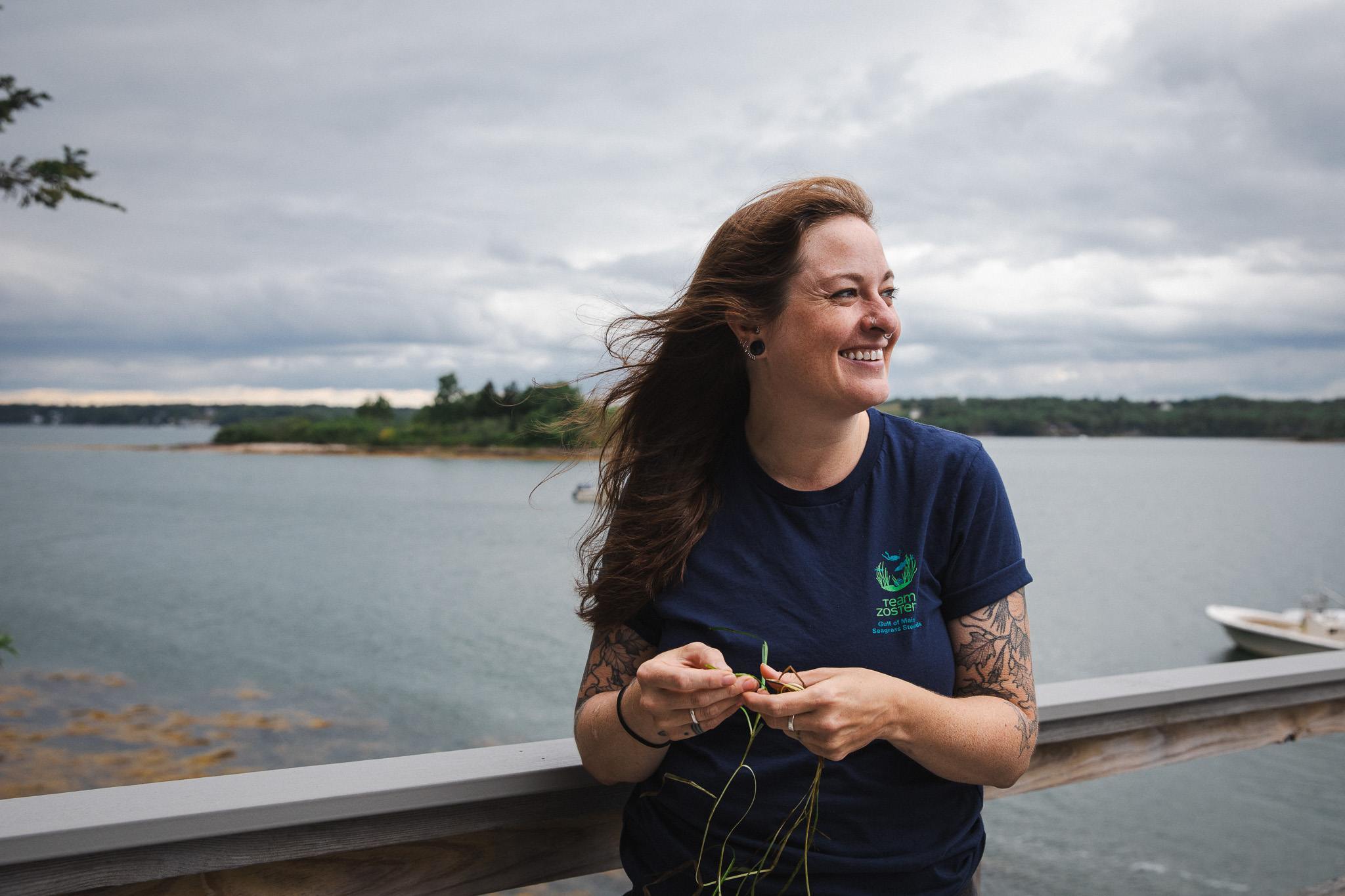 Allison Fogg smiles while holding strands of eelgrass, wearing a Team Zostera shirt, with the water and a distant boat behind her.