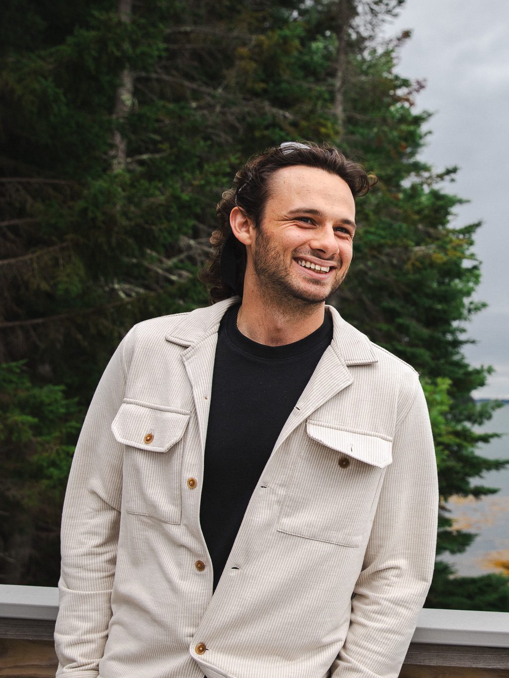 Luc Harrison smiles while standing on a dock overlooking the water at the Schiller Coastal Studies Center, with pine trees and a rocky, seaweed-covered shoreline in the background.
