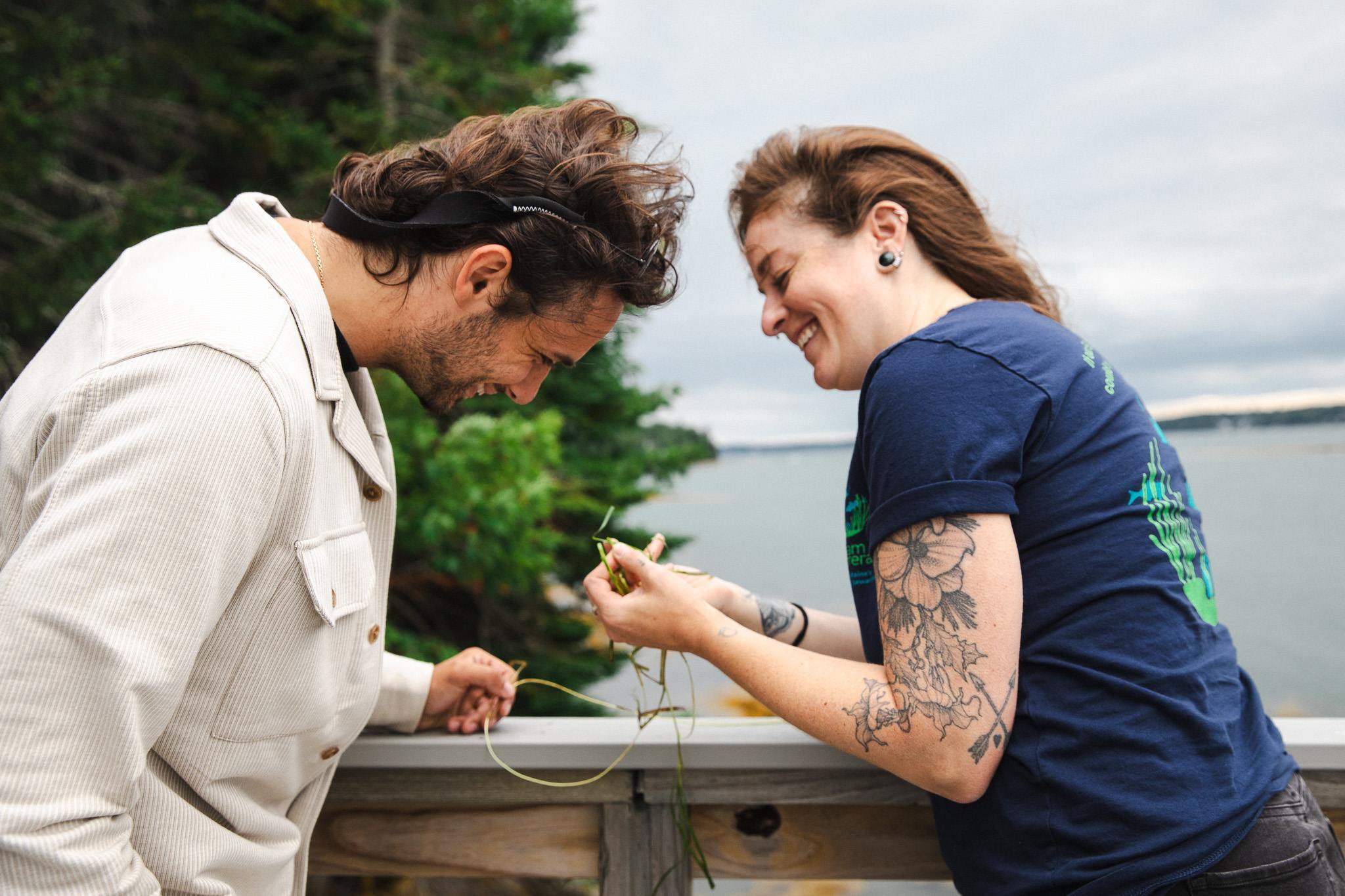 Luc Harrison and Allison Fogg laugh together while examining strands of eelgrass, leaning over a railing with the water behind them.