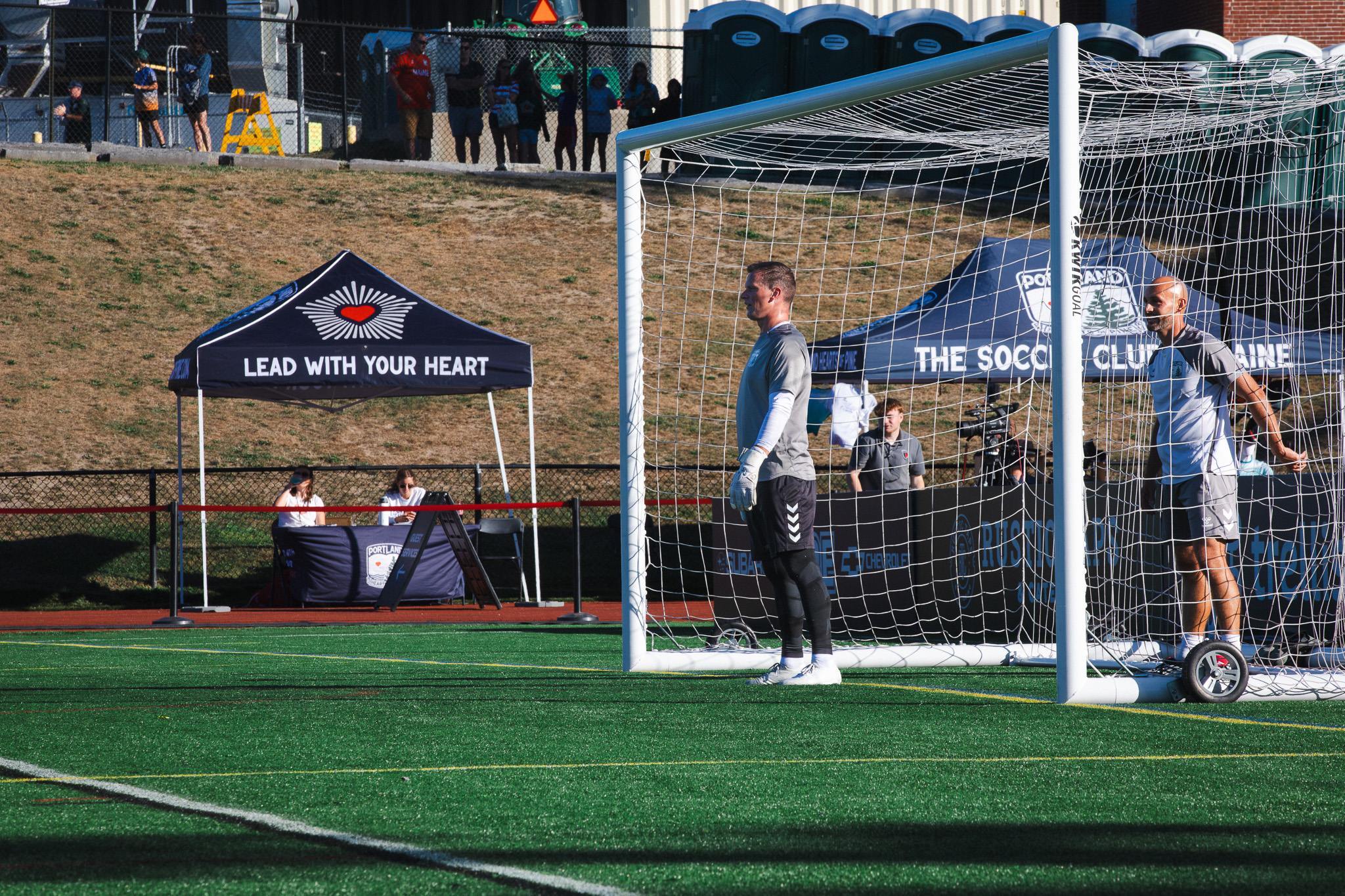 Camden Clark, wearing a Portland Hearts of Pine training kit, stands in the goal beside a keeper on the field at Fitzpatrick Stadium.