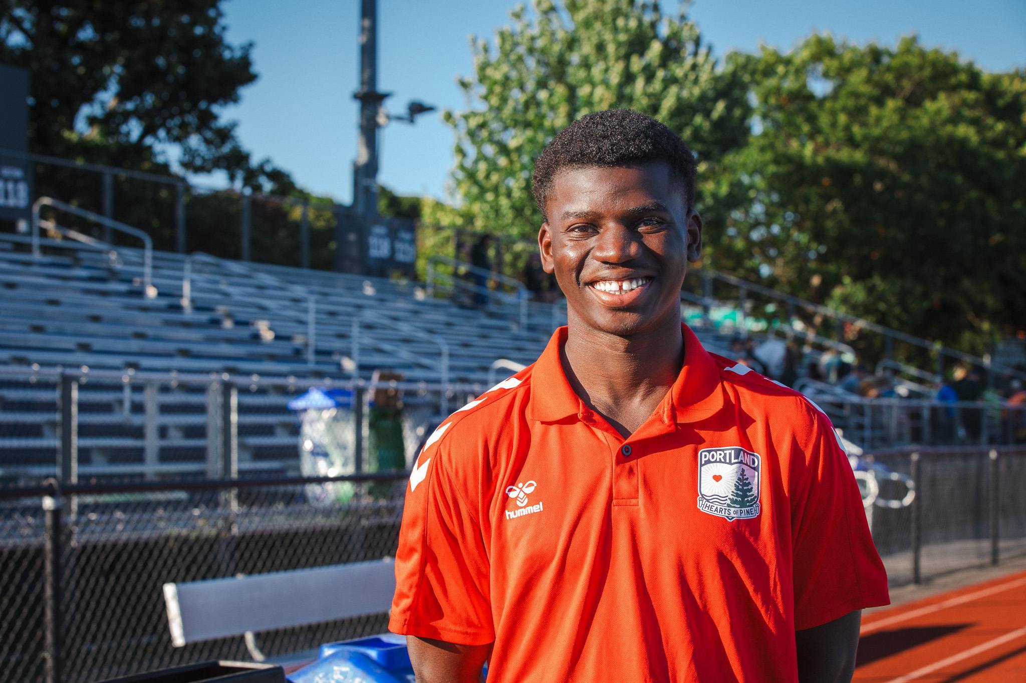 Paul Liwanga, wearing a Portland Hearts of Pine polo, smiles in the stands at Fitzpatrick Stadium.
