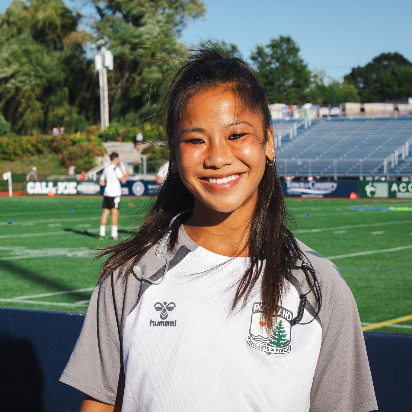 Kiaya Gatchell, wearing a Portland Hearts of Pine shirt and headset, smiles on the field at Fitzpatrick Stadium.