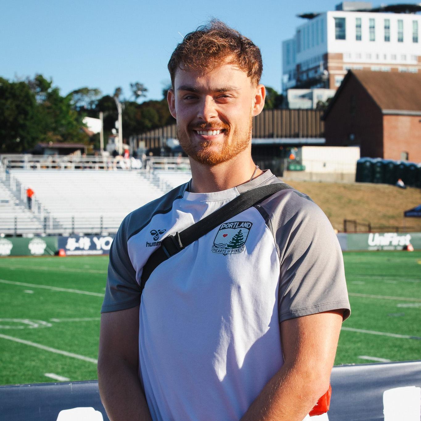 Ross Maiden, wearing a Portland Hearts of Pine shirt with a whistle around his neck, stands on the sideline at Fitzpatrick Stadium.