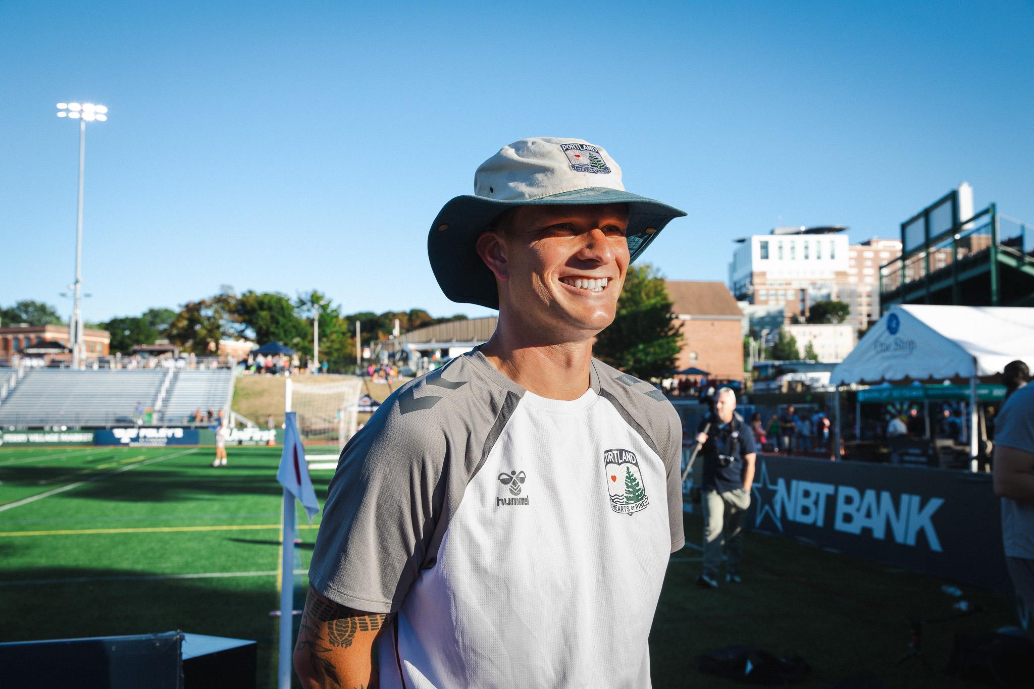 Camden Clark, wearing a Portland Hearts of Pine shirt and hat, smiles on the sideline at Fitzpatrick Stadium.
