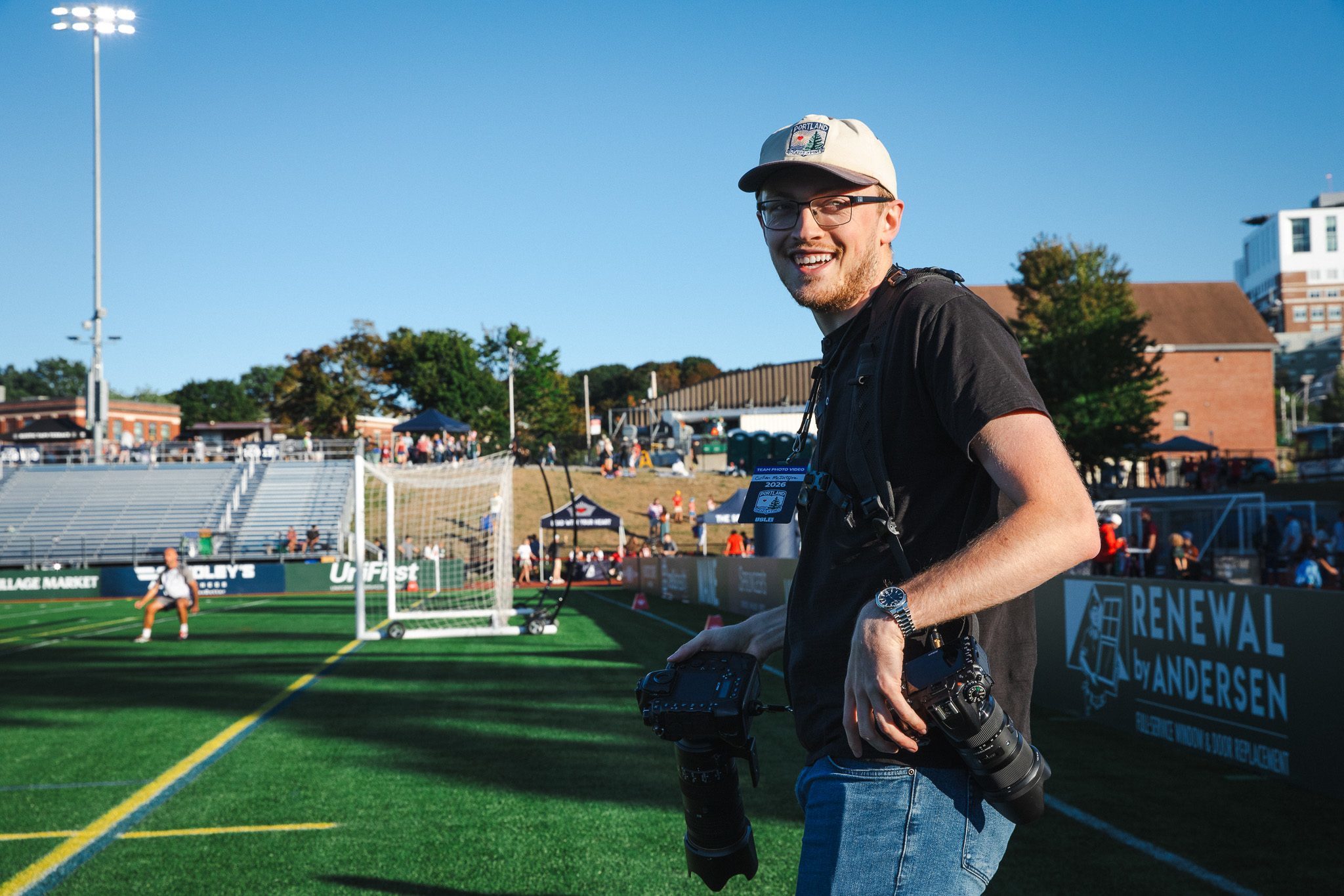 Cullen McIntyre, wearing a Portland Hearts of Pine cap and holding two cameras, smiles on the sideline at Fitzpatrick Stadium during a match.