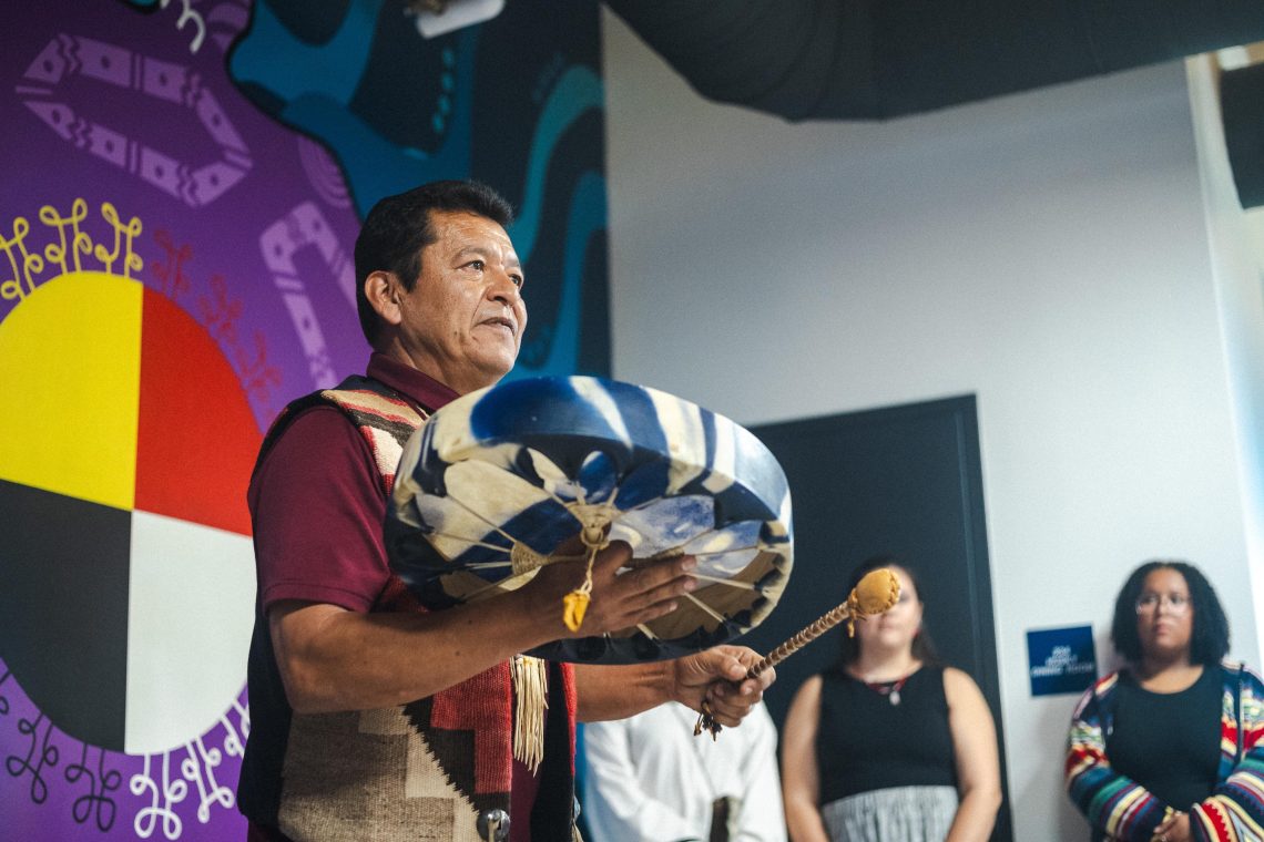 A performer plays a hand drum during USM's Indigenous Peoples' Day Celebration, with a medicine wheel mural in the background.