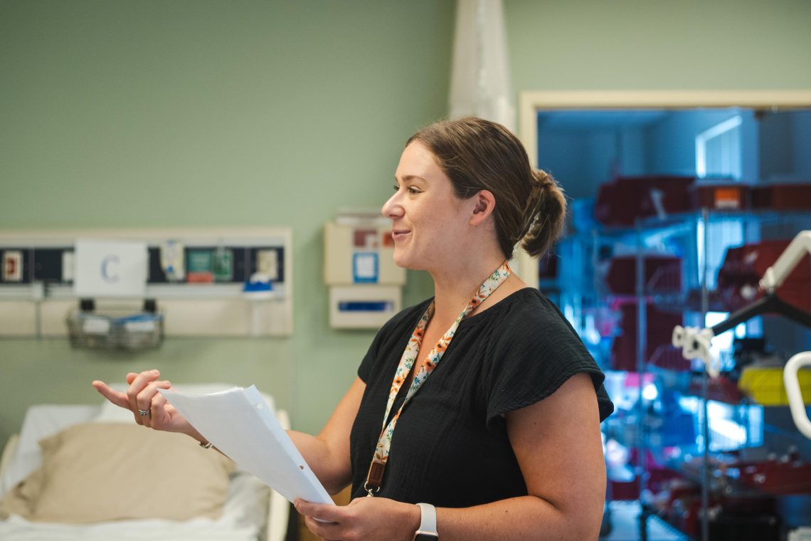A woman with a floral lanyard speaks and gestures while holding papers, standing in a nursing simulation lab beside a hospital bed and medical equipment.
