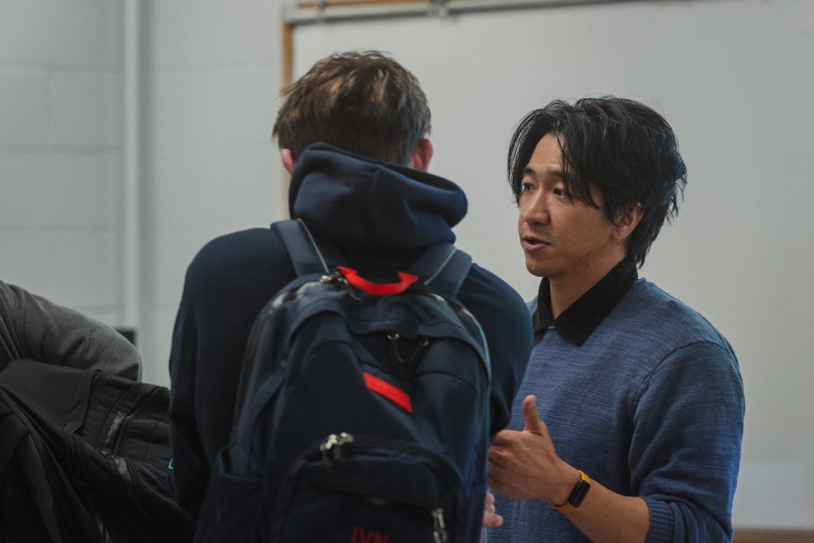 A man gestures while talking to another person wearing a backpack, in a classroom setting.