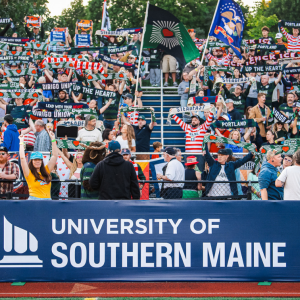 Hearts of Pine fans wave scarves and flags behind a University of Southern Maine banner at Fitzpatrick Stadium.
