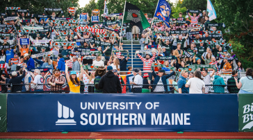Hearts of Pine fans wave scarves and flags behind a University of Southern Maine banner at Fitzpatrick Stadium.
