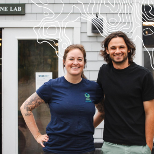 Allison Fogg and Luc Harrison smile together outside the Marine Lab at Schiller Coastal Studies Center, with a white topographic map overlay.