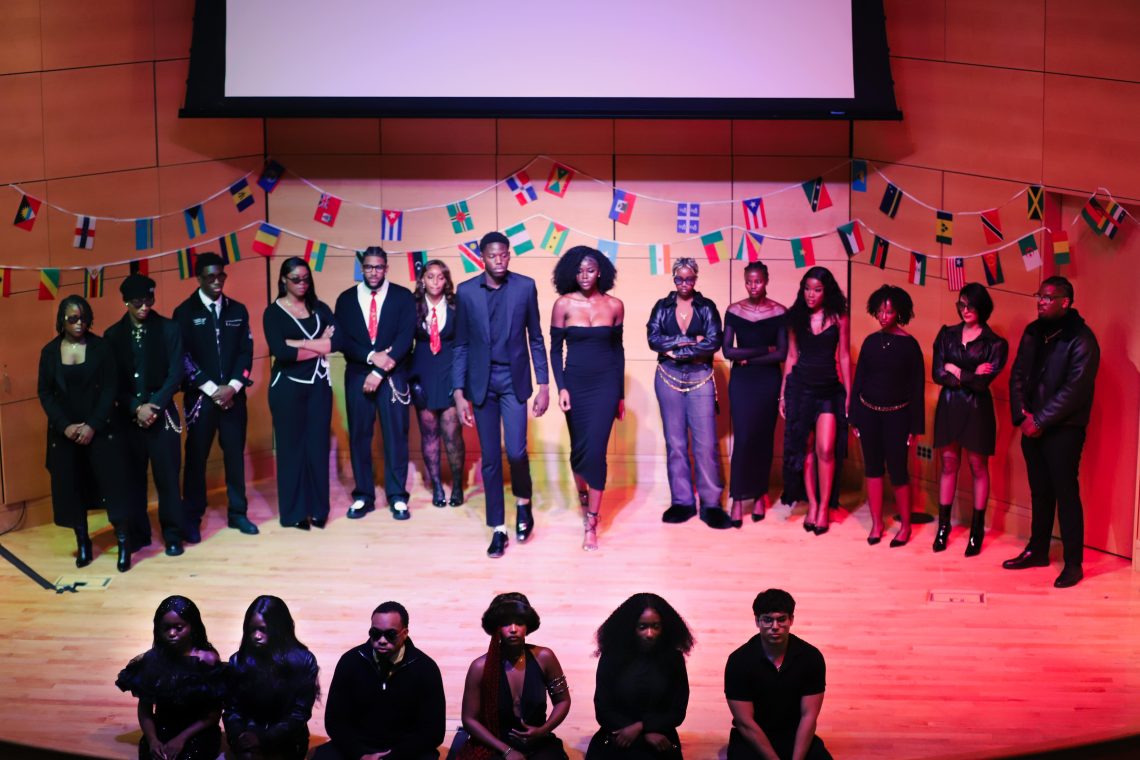A large group of USM students pose on stage in all-black fashion under a banner of international flags at Maine's Afrochella.