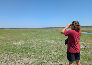 Calvin Soule looks through binoculars across Collins Marsh in Wisconsin on a clear, sunny day, with a vast expanse of wetland and cracked dry earth stretching toward a tree line in the distance.