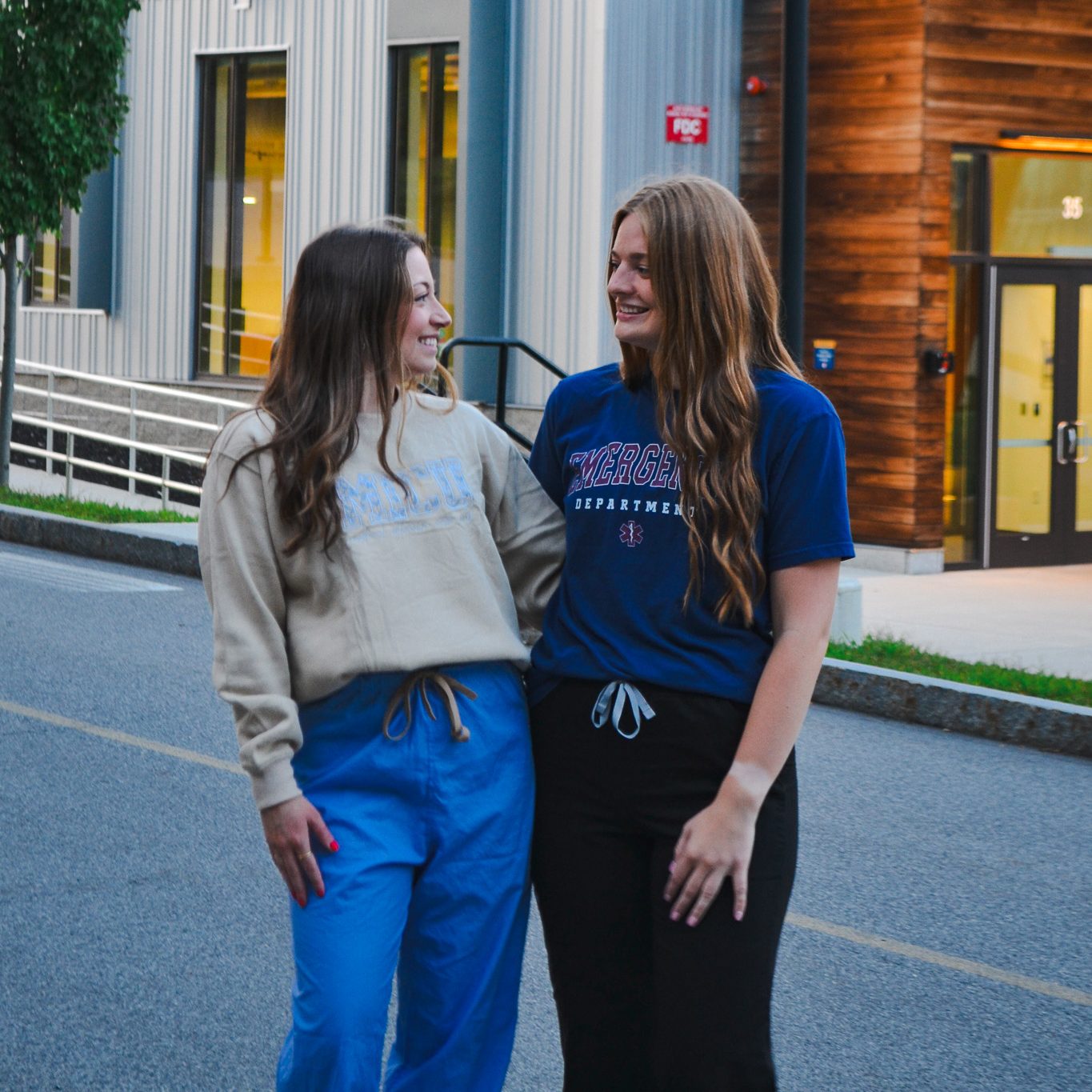 Jaden Dalrymple and her classmate smile at each other as they stand arm in arm on a campus road outside a University of Southern Maine building. One wears a tan sweatshirt and blue scrub pants, the other a navy "Emergency Department" t-shirt and black pants.