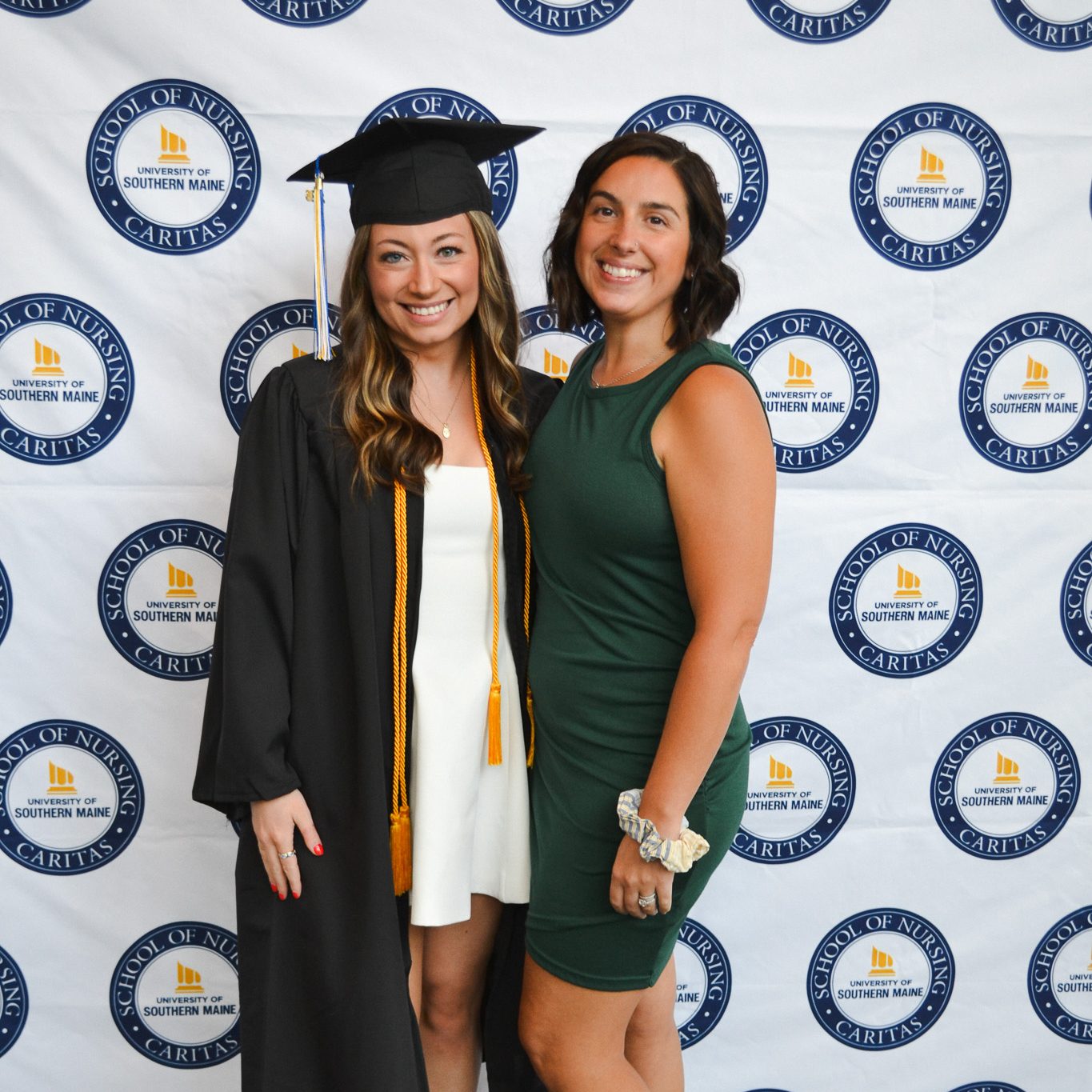 Jaden Dalrymple, in a black cap and gown with a gold honor cord, poses with her professor Britney Meunier in front of a University of Southern Maine School of Nursing backdrop.