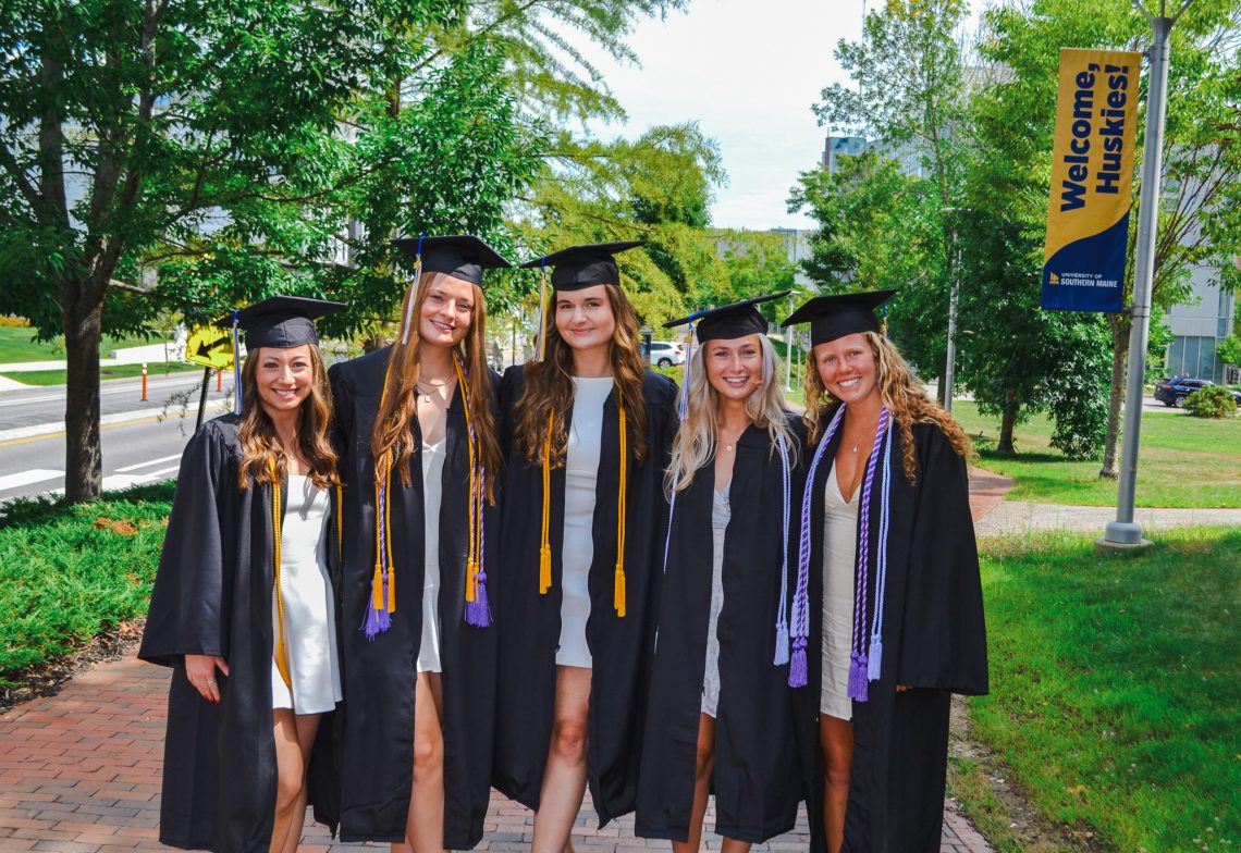 Jaden Dalrymple, and four other USM nursing graduates in caps and gowns smile together on campus. A "Welcome, Huskies!" banner hangs on the lamppost behind them.