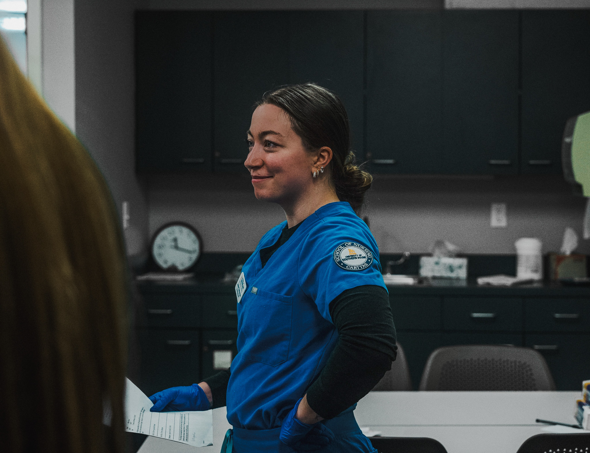 Jaden Dalrymple, a USM nursing graduate, smiles in blue School of Nursing scrubs and gloves, standing in a nursing lab with one hand on her hip.
