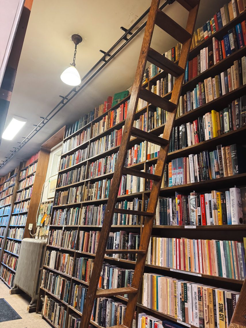A wooden rolling ladder leans against floor-to-ceiling shelves packed with books in a Portland bookstore, lit by a vintage pendant lamp.