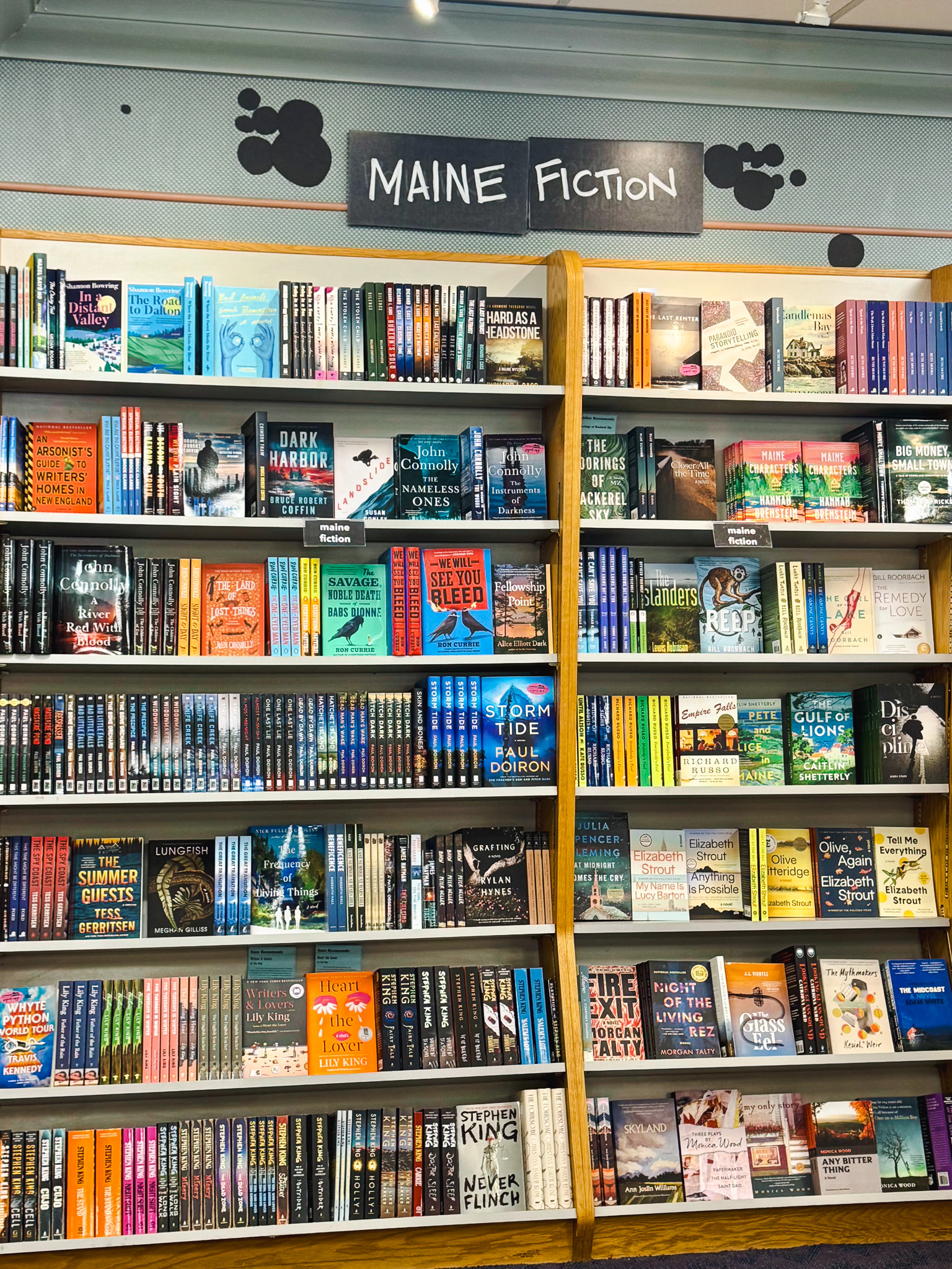 A tall bookcase under a “Maine Fiction” sign, filled with novels by Maine authors including Stephen King, Elizabeth Strout, Richard Russo and Paul Doiron.