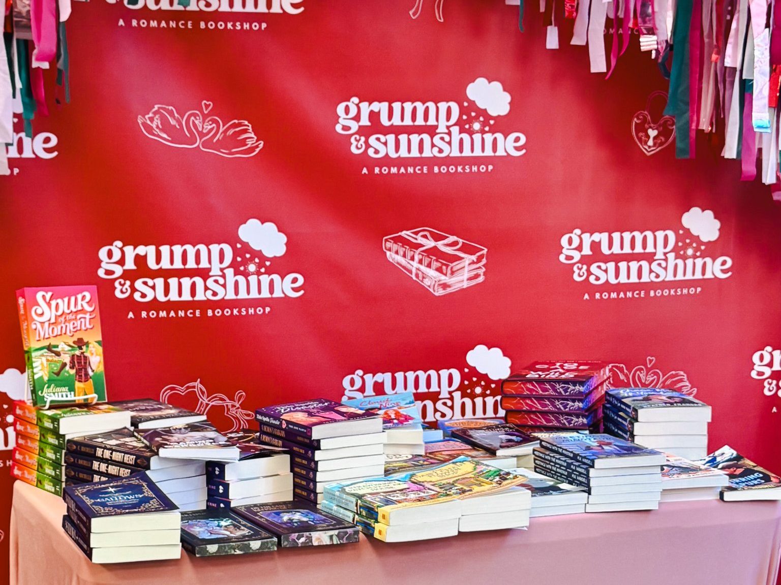 A table of stacked romance novels in front of a red Grump & Sunshine romance bookshop backdrop, framed by hanging pink, red, white and green streamers.