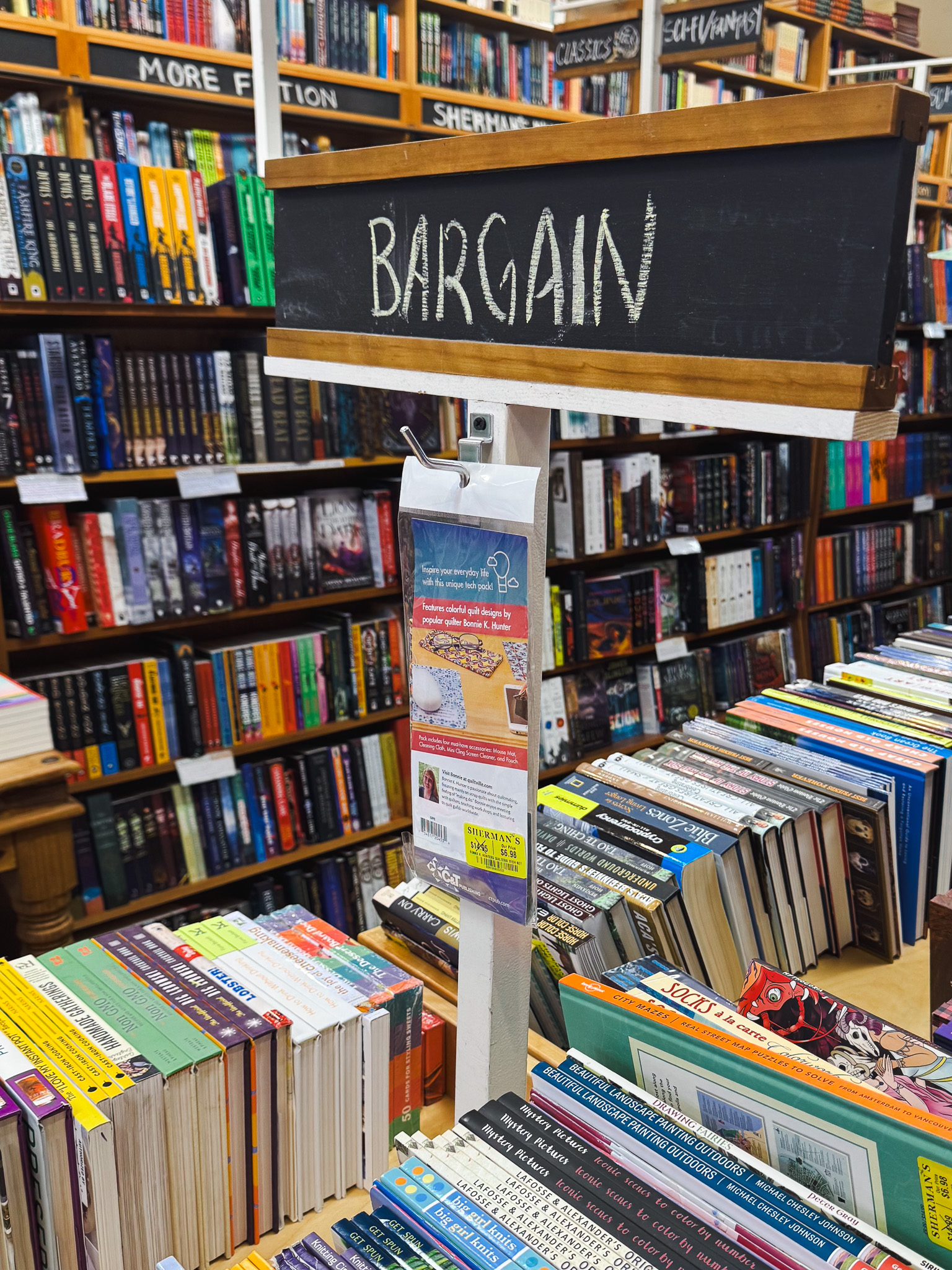 A chalkboard sign reading “Bargain” stands over tables of discounted books at Sherman’s, with packed shelves labeled “More Fiction,” “Classics” and “Sci-Fi/Fantasy” in the background.