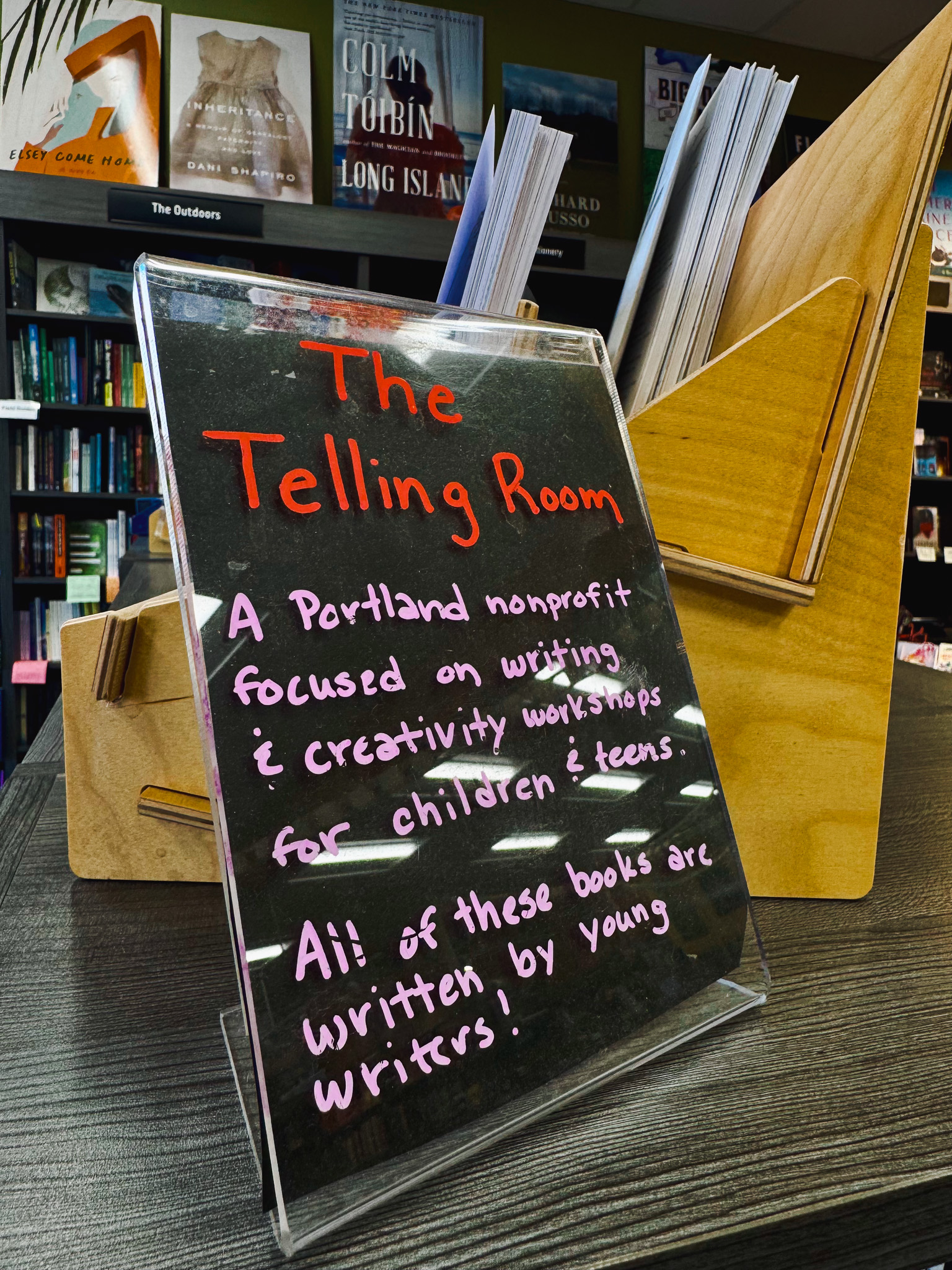 A handwritten sign in a bookstore reads, “The Telling Room: A Portland nonprofit focused on writing and creativity workshops for children and teens. All of these books are written by young writers!” A wooden display of the books stands behind it.