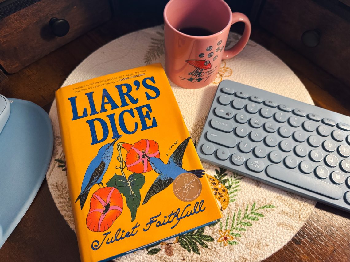 A hardcover copy of “Liar’s Dice” by Juliet Faithfull, with a yellow cover showing blue hummingbirds and orange flowers, sits on a woven placemat beside a pink coffee mug and a light blue keyboard.