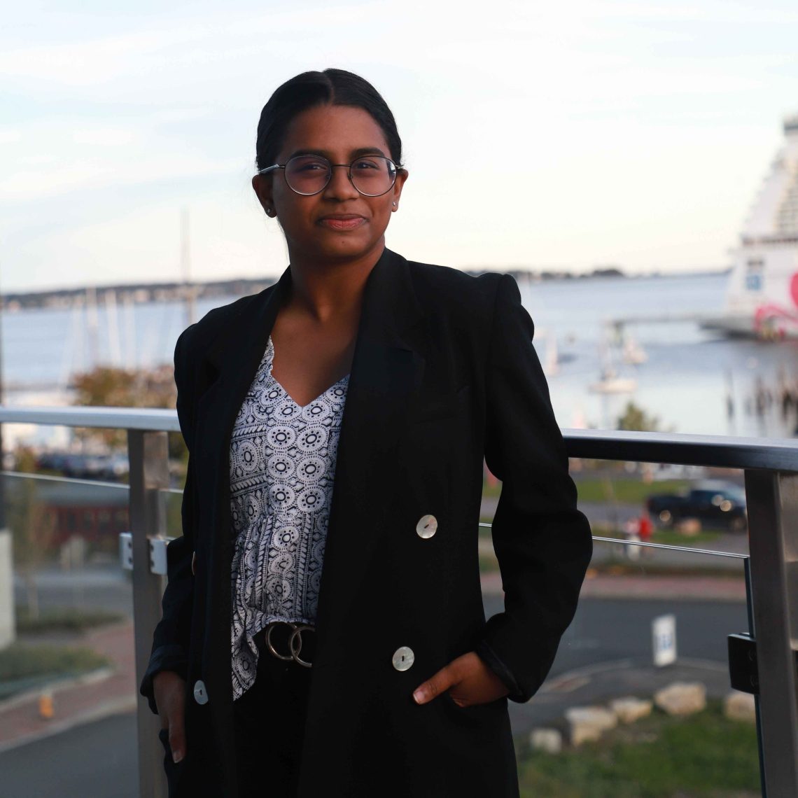 Vanathi Selvan stands on a balcony in a black blazer and glasses, with a harbor and boats behind her.
