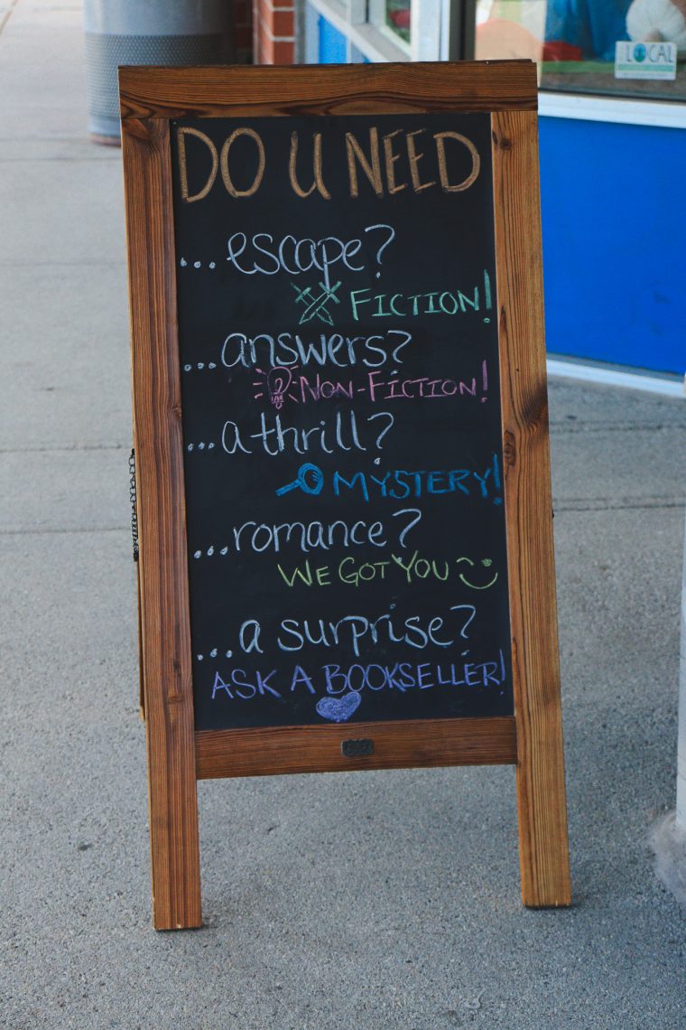 A wooden sidewalk chalkboard sign outside a bookstore reads, “Do U Need... escape? Fiction! ...answers? Non-fiction! ...a thrill? Mystery! ...romance? We got you. ...a surprise? Ask a bookseller!”