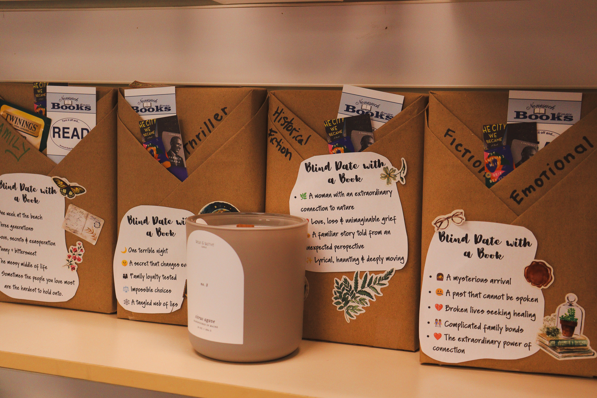A row of books wrapped in brown kraft paper, each labeled “Blind Date with a Book” with a genre and a short list of teaser clues, with bookmarks and a tea bag tucked into the wrapping beside a candle.