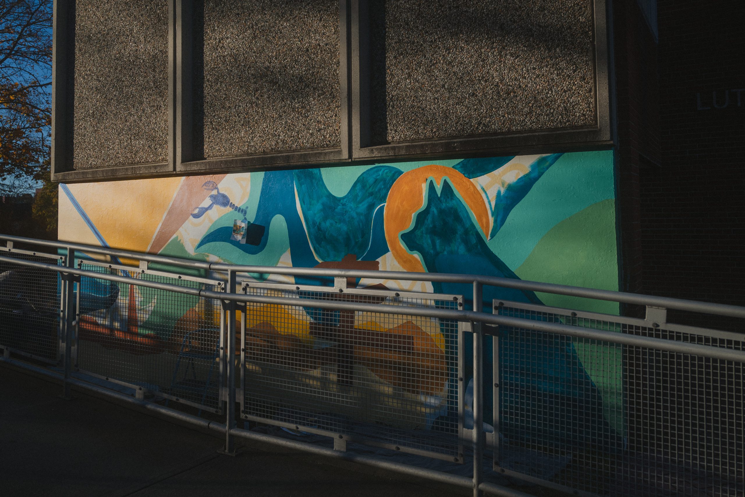 The in-progress Southern Maine mural on Luther Bonney Hall, with a blue lobster and a husky silhouette against an orange sun, seen through a metal railing in low afternoon light.