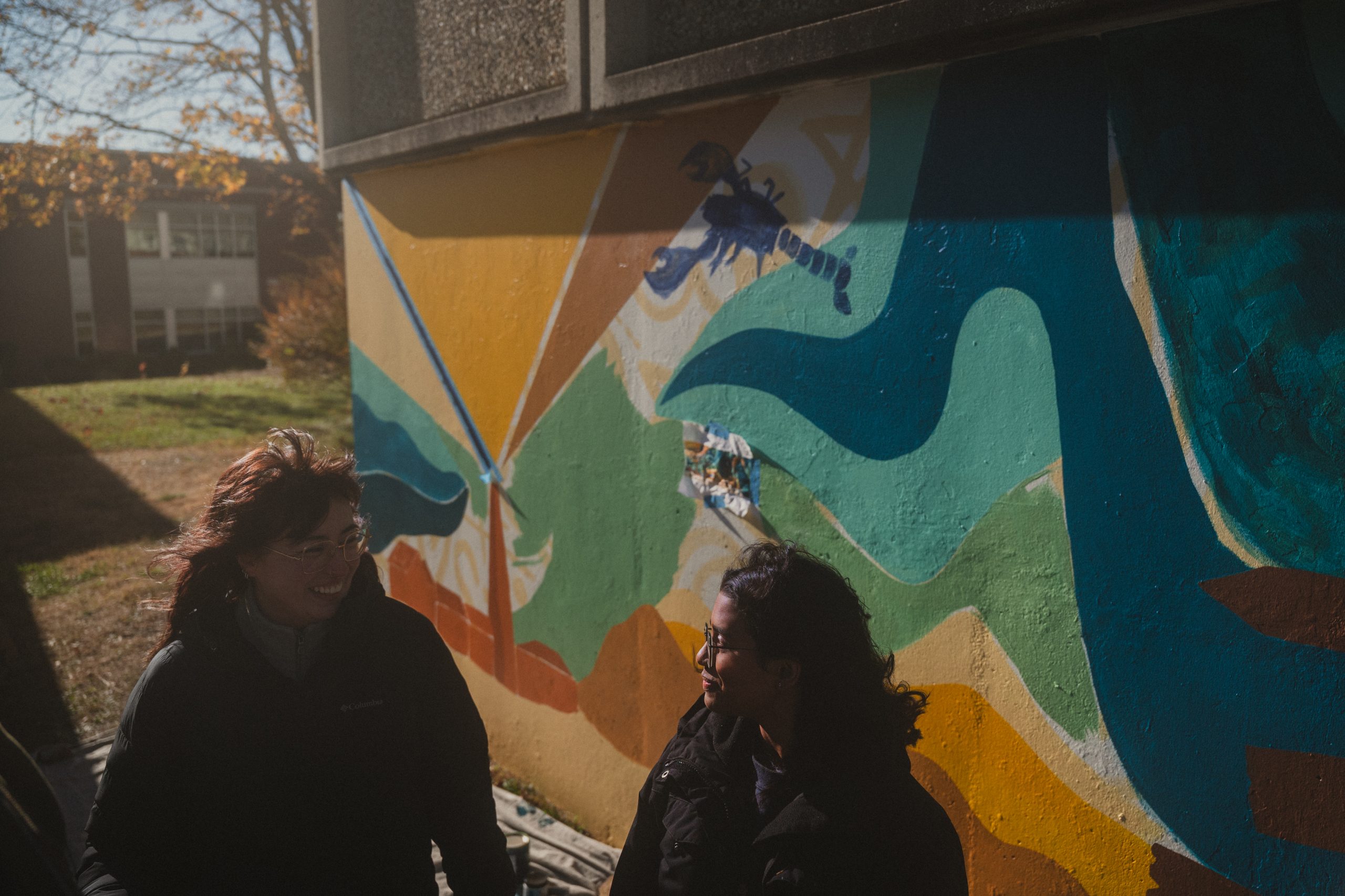 Two artists laugh in the afternoon sun in front of their in-progress Southern Maine mural at USM, with a blue lobster painted behind them.
