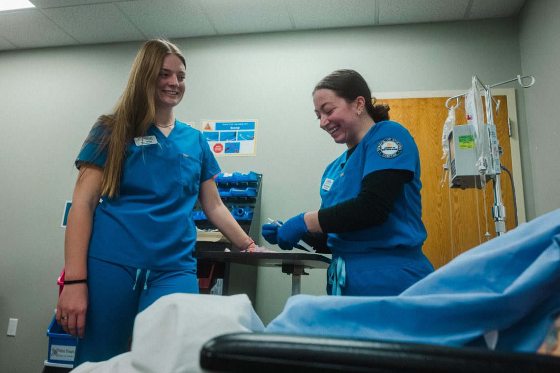 Jaden Dalrymple (right), a USM nursing student in blue scrubs and gloves, laughs while preparing a syringe in a simulation lab as a classmate looks on.