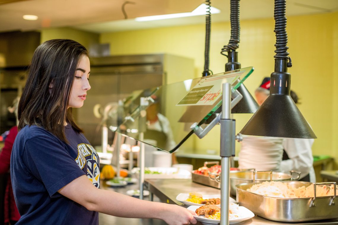 A student in a USM Huskies T-shirt picks up a plate of food from a serving line at Brooks Dining Hall on the Gorham campus.