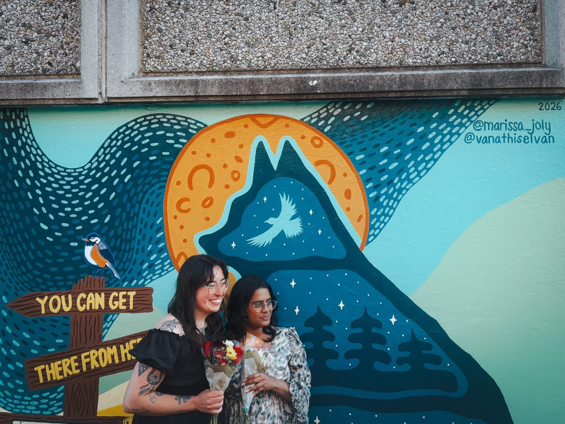 The two artists smile at the mural unveiling, each holding a bouquet, in front of the husky silhouette, golden sun, and “You Can Get There From Here” signpost.