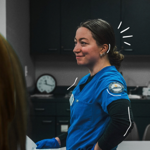 Jaden Dalrymple, a USM nursing graduate in blue School of Nursing scrubs and gloves, smiles and looks off to the side in a nursing lab.
